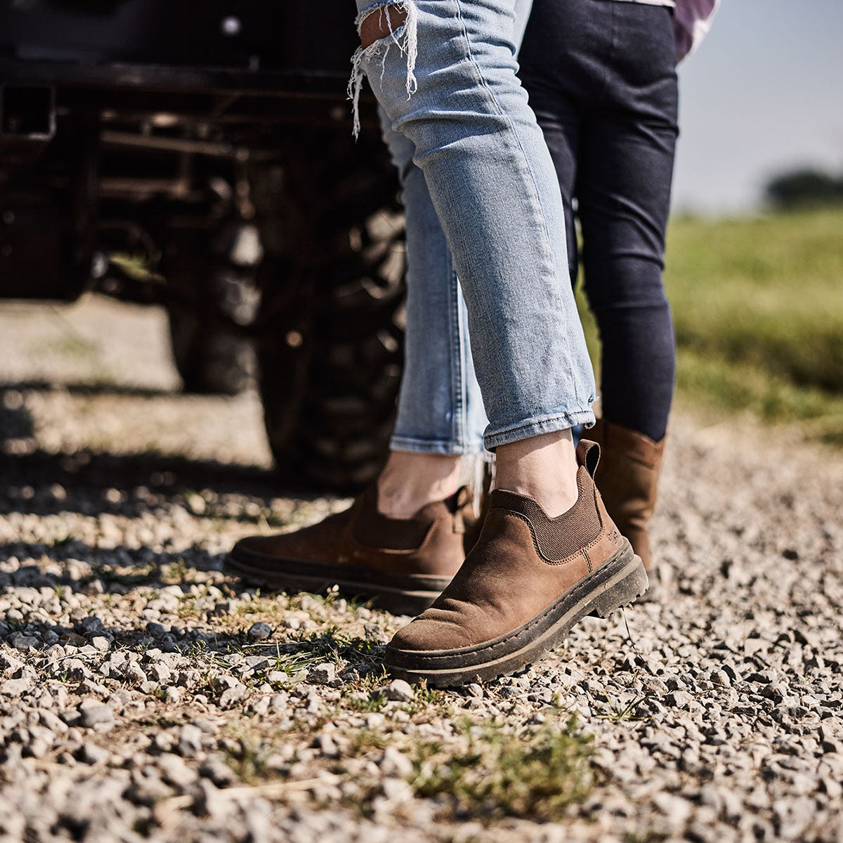 Two people wearing brown boots walking on a gravel path with a vehicle in the background.