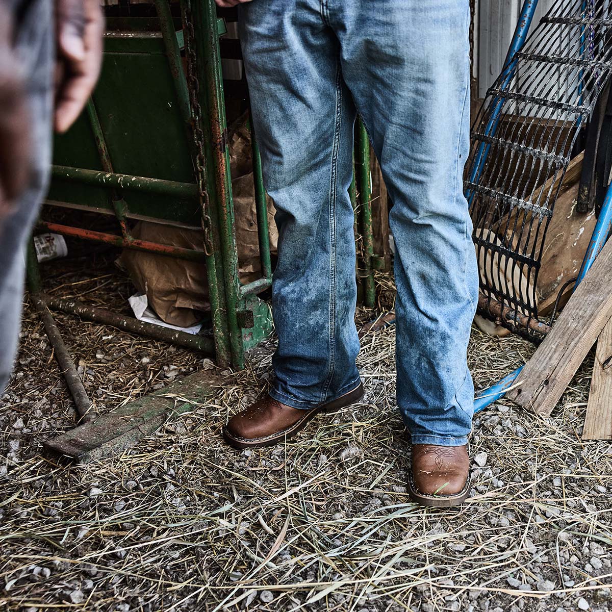 Person wearing blue jeans and brown boots standing on a dirt floor with tools and equipment in the background.